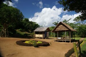 Big clouds billow over a neatly swept yard surrounded by green trees. Two bamboo huts are in the yard; one is open-sided and has chairs and a table.