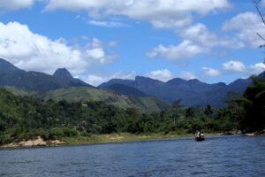 A canoe bobs on a wide, blue river lined with green trees.